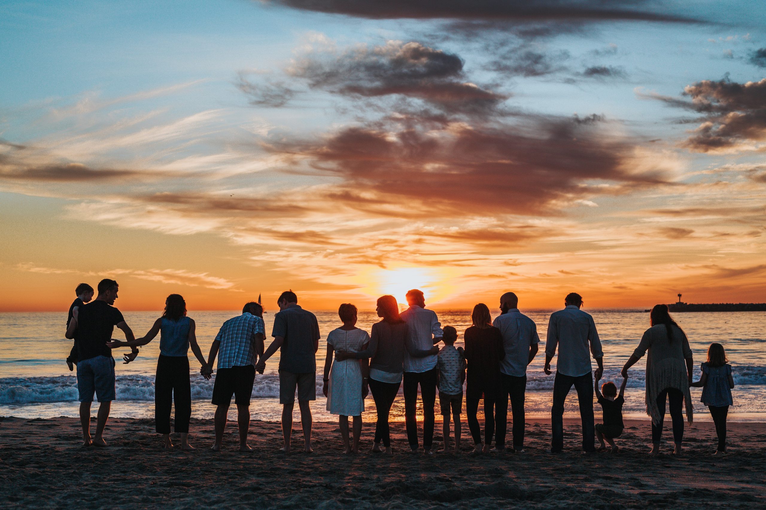 una fila di persone che guardano il tramonto al mare, tenendosi per mano o abbracciati, fra loro anche dei bambini