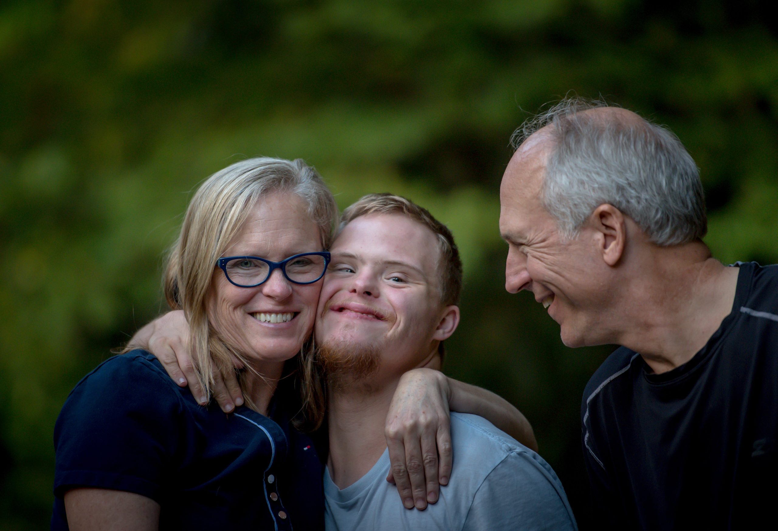 mamma abbracciata al figlio con la sindrome di down che sorride, a fianco al padre, sorridente anche lui.